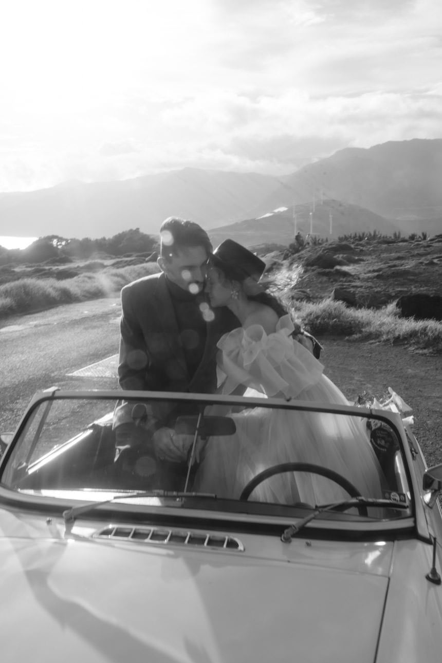 Bride and groom sitting in the backseat of a convertible, with a hat blowing in the wind, capturing a carefree wedding moment.