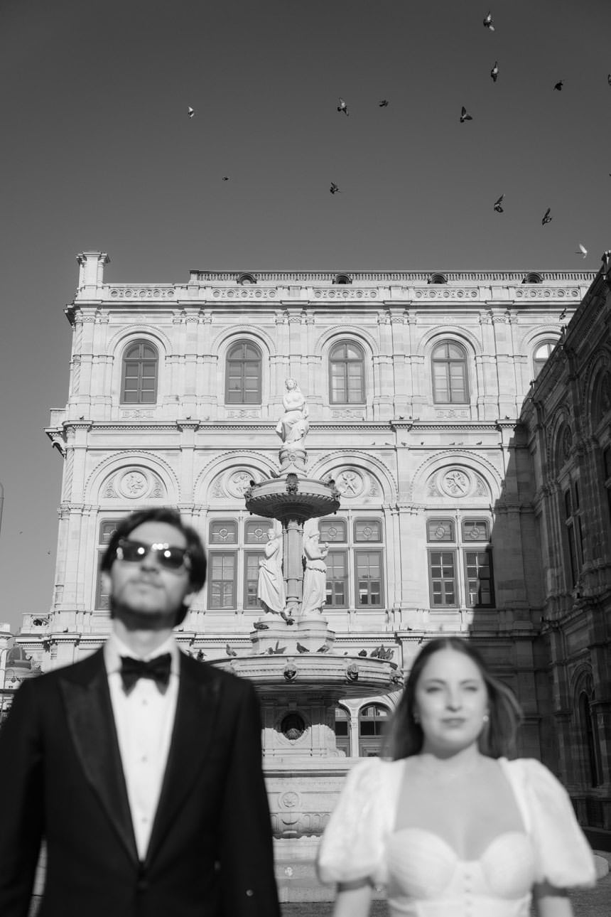 Bride and groom standing side by side in front of the Vienna Opera House as birds fly past, creating a cinematic wedding moment.