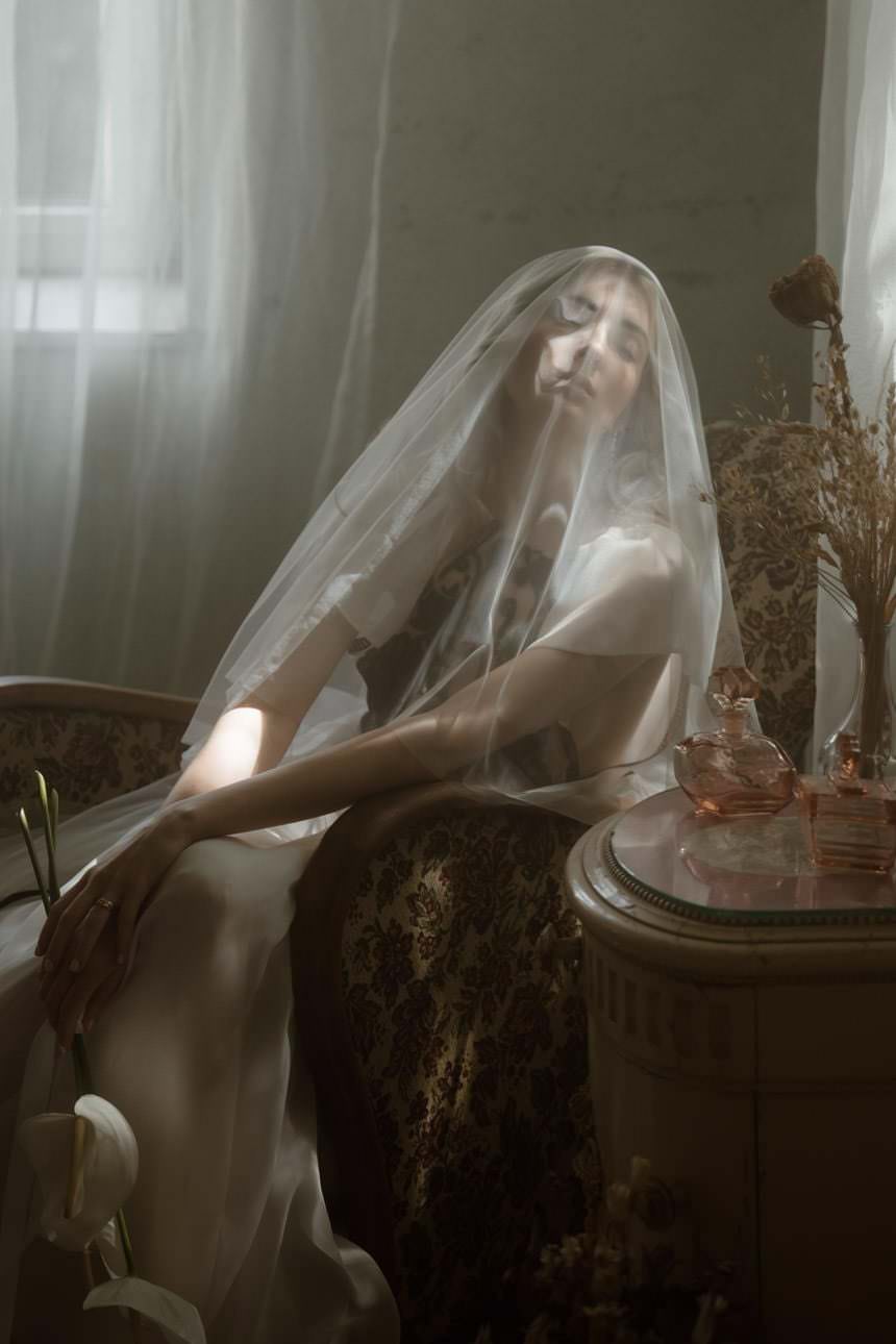 Bride sitting on an antique chair in a historic room, with a veil over her face and holding a flower in her hand.
