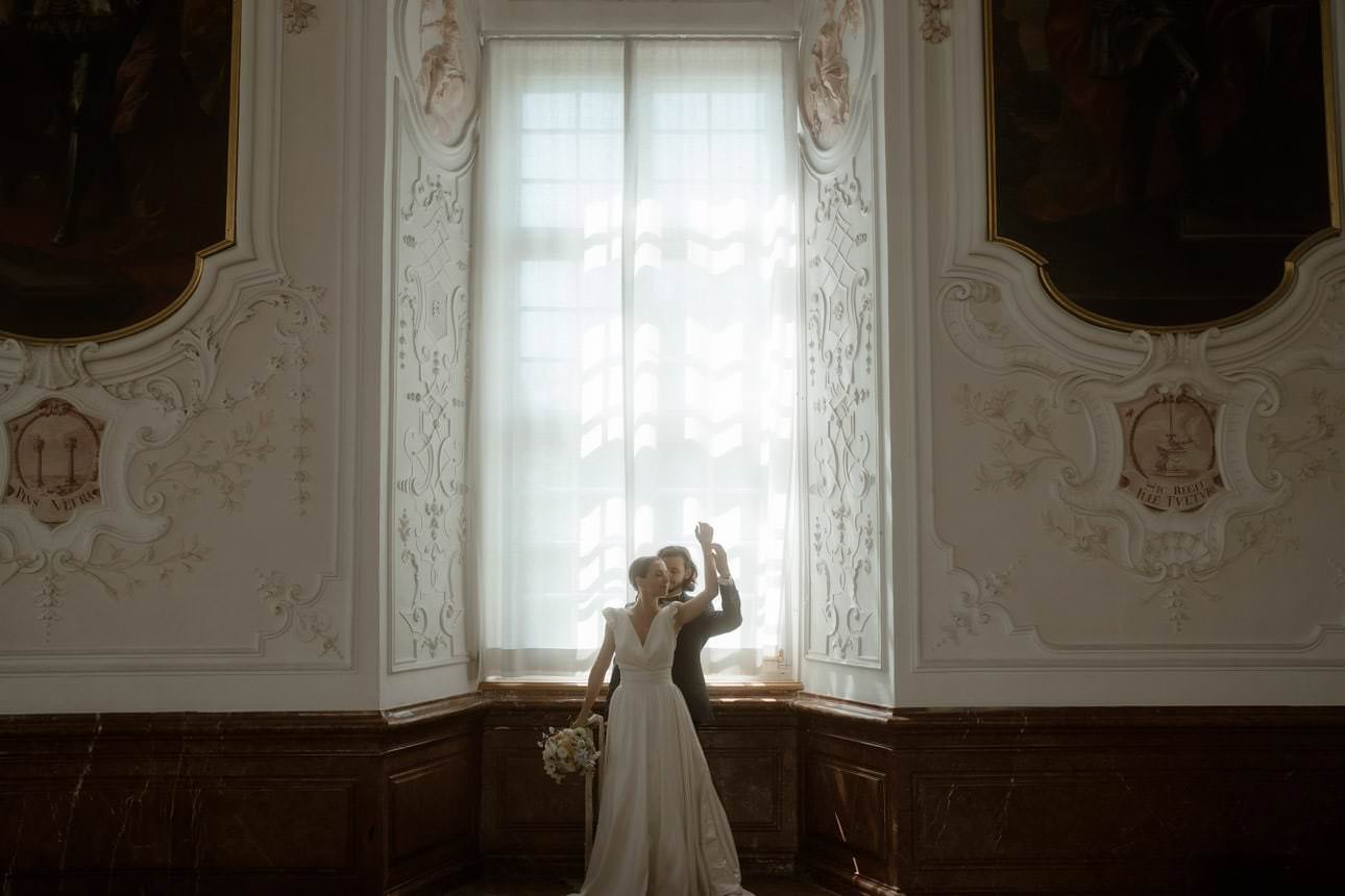 Groom standing behind his bride, gently stroking her arm by a window in a luxurious historic location.