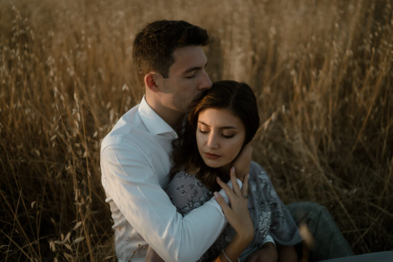 At sunrise in a field near Agriturismo Baccoleno, a couple sits in the tall grass—he kisses her head gently as she rests her hand on his arm, capturing an intimate moment during their Tuscany photo shoot. Sunrise Couple Photo Shoot Tuscany Agriturismo Baccoleno Pienza.